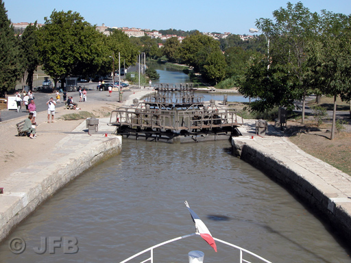 Une péniche, dont on ne voit que la proue, descend vers béziers. On aperçoit en perspective les trois derniers vantaux de l'écluse. C'est l'été, les arbres sont verts, et l'on aperçoit des touristes sur les berges.