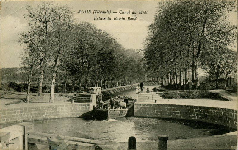 Carte postale ancienne légendée : Agde (Hérault) - Canal du Midi - Écluse du bassin rond. On aperçoit une péniche en train de franchir la vanne en direction de Sète. Au tout premier plan, on aperçoit le vanne fermée en direction de Béziers. La forme ronde du bassin est nettement visible.