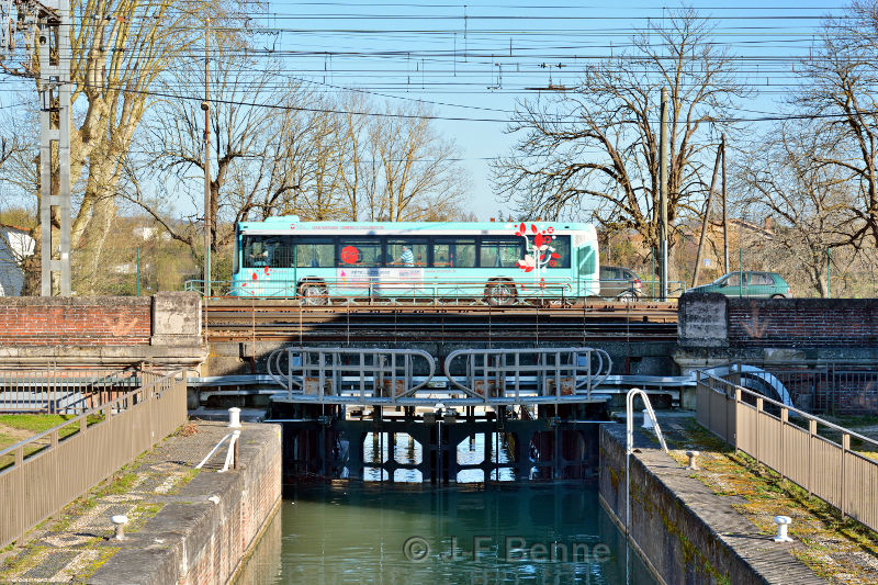 Une photo de l'écluse de Montauban, sur le Canal de Montech, sur laquelle passe aussi la route et la voie ferrée. On voit un bus de la ville peint en vert clair qui passe sur l'écluse.