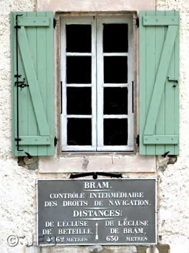 Une fenêtre avec des volets verts, et au dessous, le panneau des distances qui indique : Bram, contrôle intermédiaire des droits de navigation<br />Distances : de l'écluse de Béteille (4962m) à l'écluse de Bram (630m).