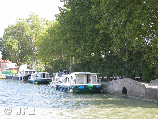 Image d'été : Trois bateaux amarrés, et à droite, un petit épanchoir, en forme de petit pont, qui permet l'écoulement des eaux excédentaires sur le Grand Bassin de Castelnaudary.