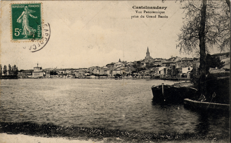 Carte postale ancienne légendée : Castelnaudary, vue panoramique prise du grand bassin. De gauche à droite, on aperçoit l'île de la    Cybèle plantée de grands arbres, l'ensemble de la ville avec l'église qui domine et devant nous, le grand bassin. Une péniche vue de face est amarrée sur la droite.