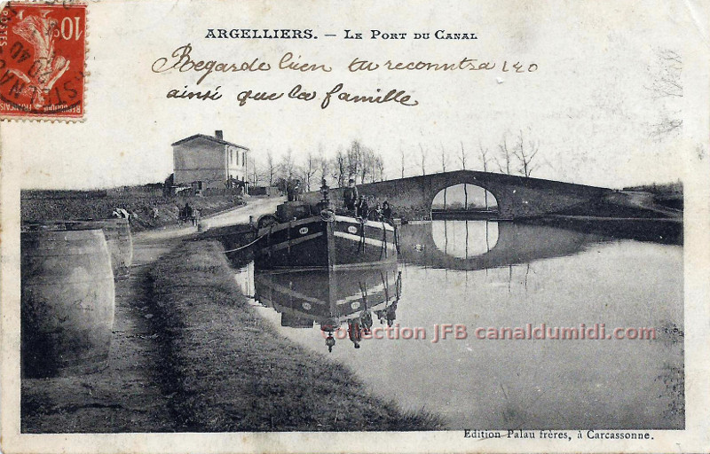 Carte postale ancienne légendée Argelliers, le port du canal. Vue du pont et de la maison du garde, et au 
premier plan, une péniche sur laquelle se trouvent quelques personnes. Il est écrit à la plume : 
Regarde bien, tu reconnaitra Léo ainsi que la famille.