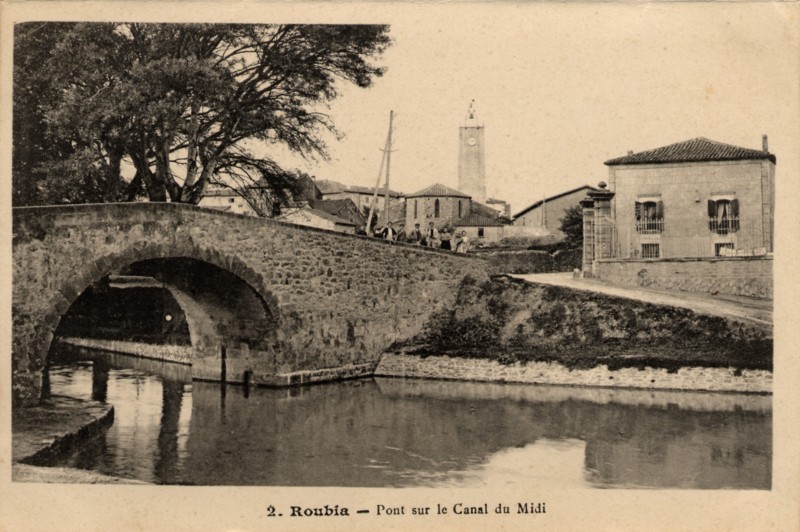 Carte Postale ancienne légendée : Roubia - Pont sur le Canal du Midi. On aperçoit aussi l'église et un petit groupe de promeneurs sur le pont.