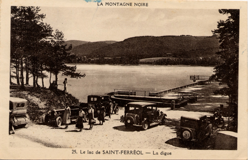 Carte postale ancienne légendée : 
La Montagne Noire - Le Lac de Saint-Ferréol
    - La Digue.  On aperçoit le lac de Saint-Ferréol à son
    niveau presque optimal, le début de la digue et le système d'évacuation
    du trop plein. Quelques tacots des années 30 sont stationnés en premier
    plan, et l'on voit quelques visiteurs vétus à la mode de l'époque
