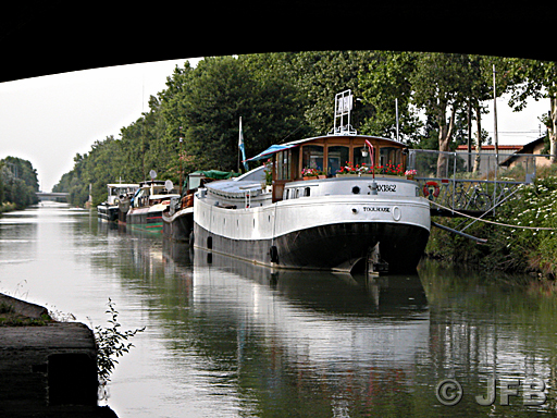 Le long du Canal de Garonne, une péniche blanche et noire dénommée Toulouse est amarrée, elle est encadrée par l'arche de l'un des ponts-jumeaux