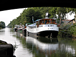 Le long du Canal de Garonne, une péniche blanche et noire dénommée Toulouse est amarrée, elle est encadrée par l'arche de l'un des ponts-jumeaux