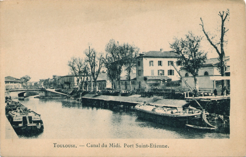 Carte postale ancienne légendée : Tousouse - Le Canal du Midi - Port Saint Étienne. On aperçoit des péniches, sur les deux rives du canal, et de l'autre côté, un lavoir couvert. De part et d'autres, des constructions qui nous montrent bien que nous sommes en ville, et au dernier plan, un pont, typique du Canal du Midi